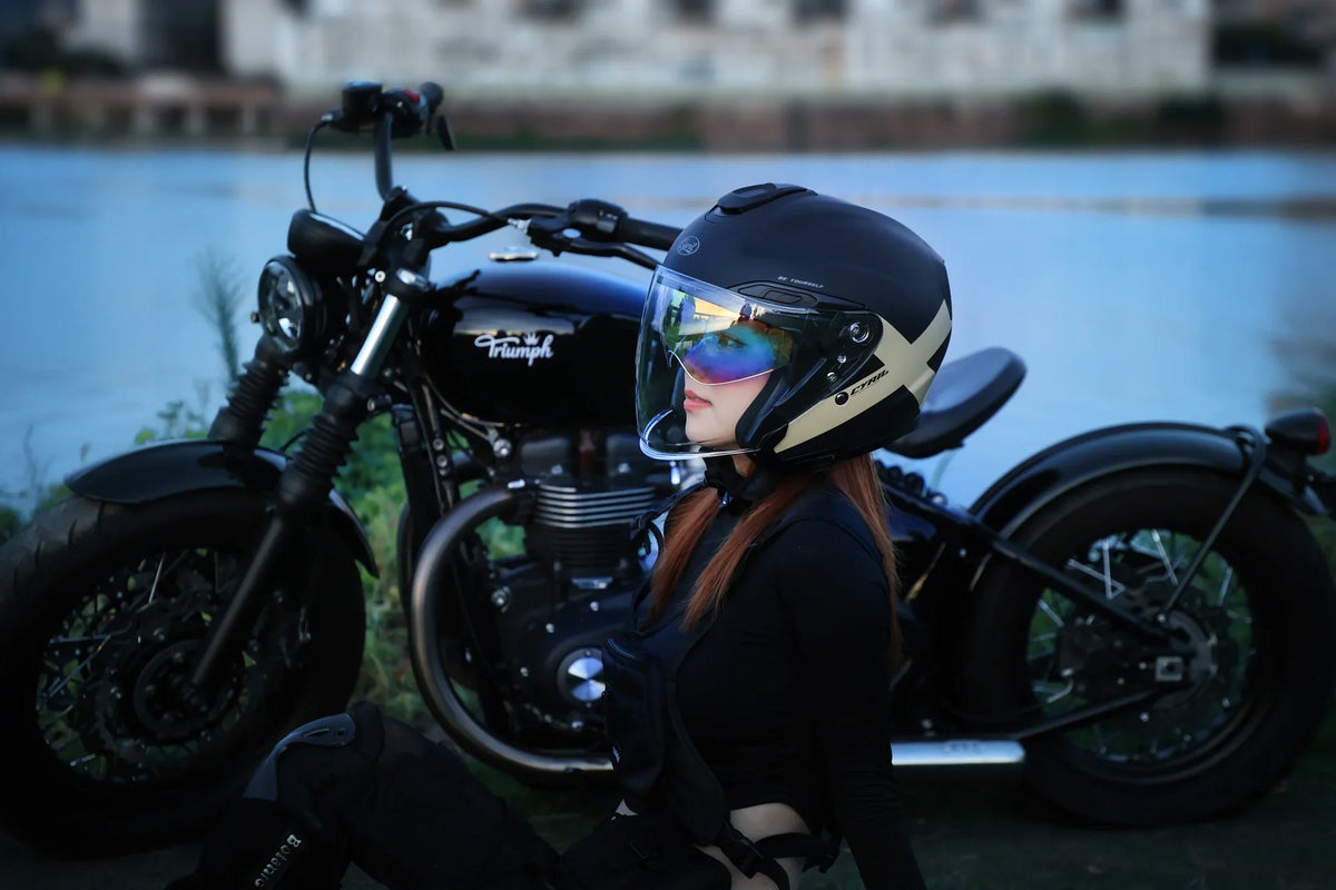 Rider in an open-face helmet and goggles standing beside a cruiser by the water