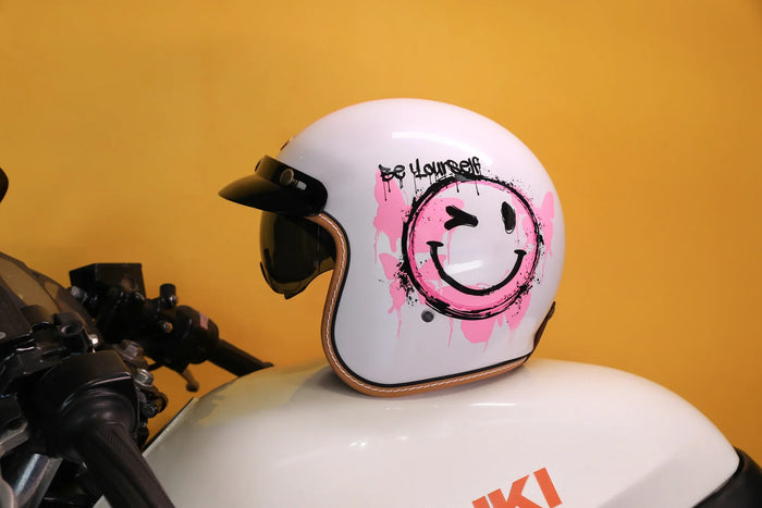 White open-face helmet with a pink smiley graphic on a motorcycle tank